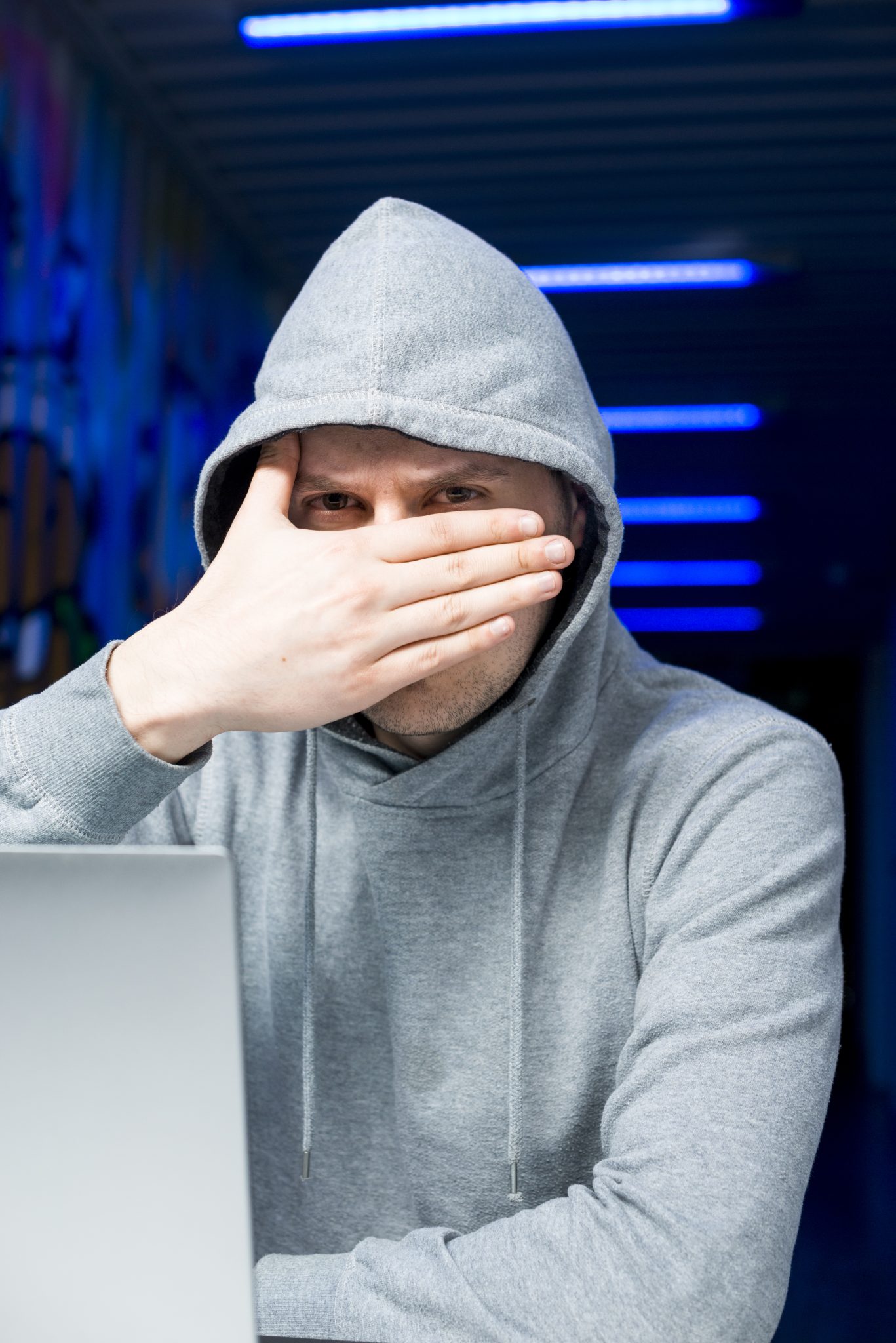 Uomo con felpa grigia e cappuccio, copre il viso con la mano davanti a un laptop.